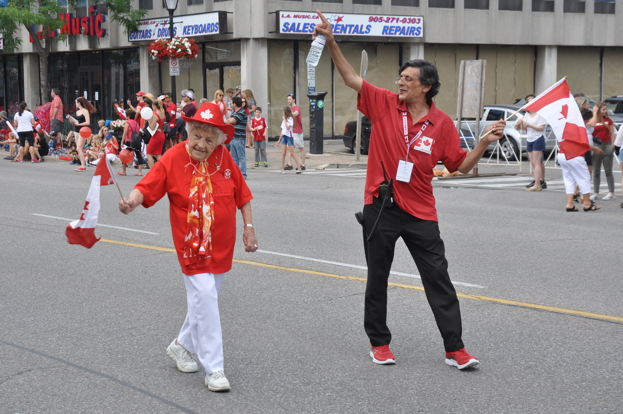 Hazel McCallion in a red maple-leaf hat, waving a Canadian flag as she walks a Canada Day parade route.